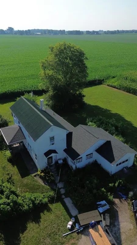 Drone view of a farmhouse roof project — dark architectural shingles alongside the green metal roof, fields behind